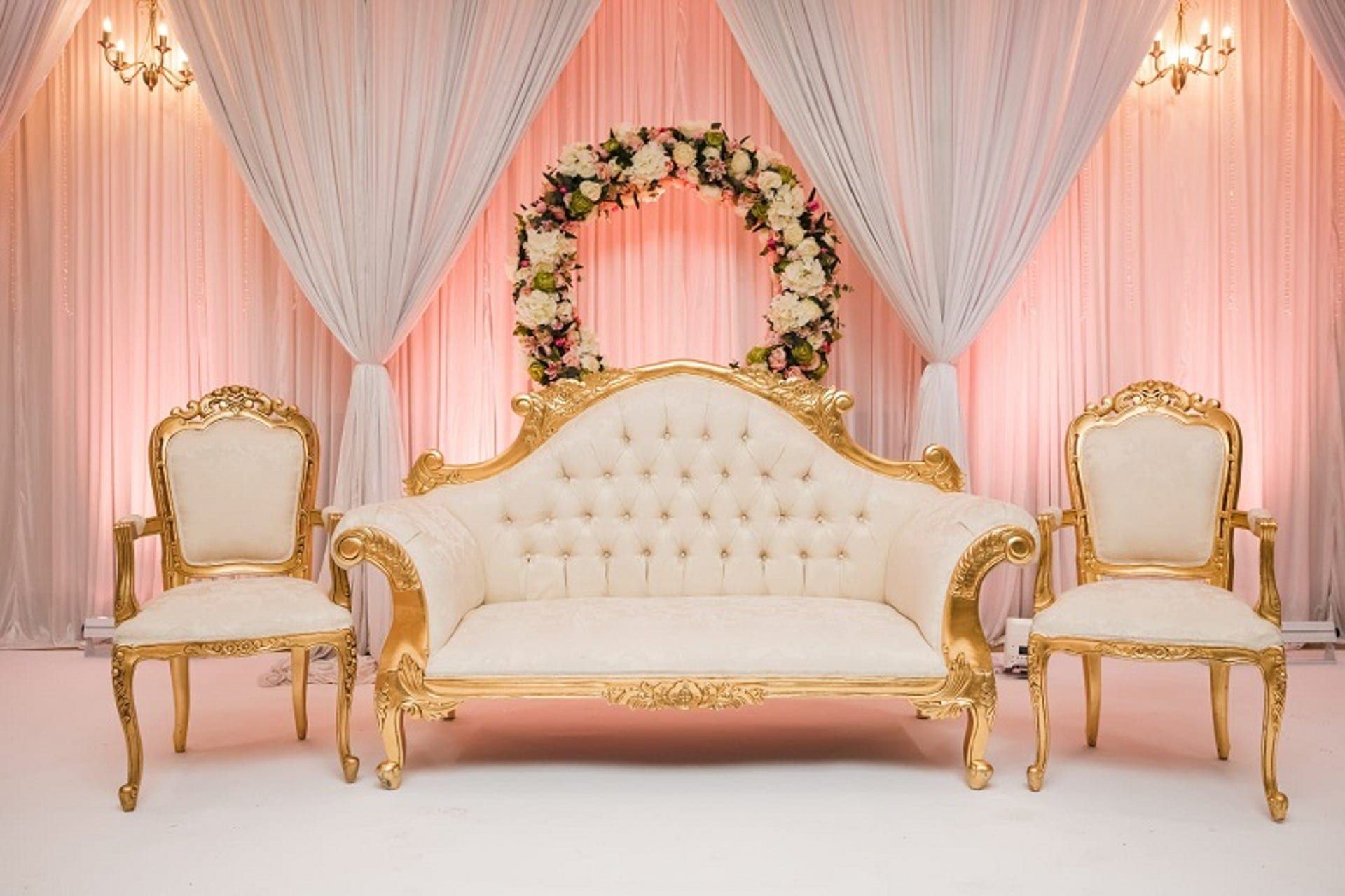 A blush draped backdrop with a floral arch behind a gold framed sofa and chairs