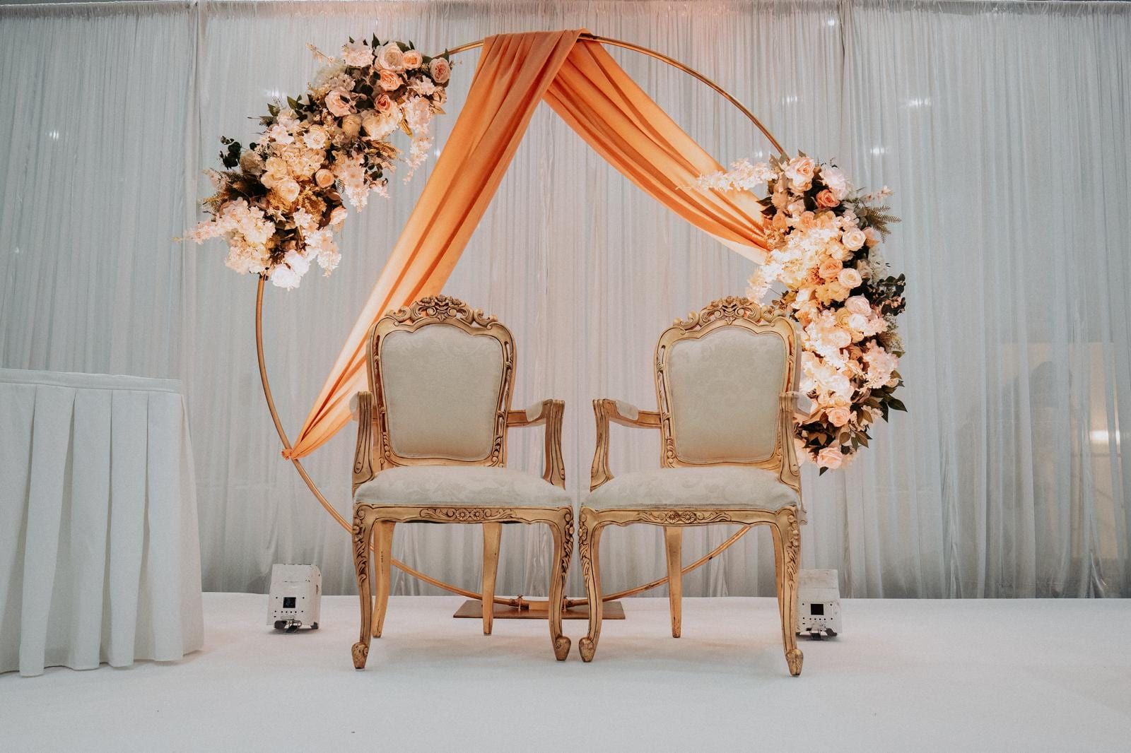A coral draped arch trimmed with blossom, framing two gilded chairs on a white stage