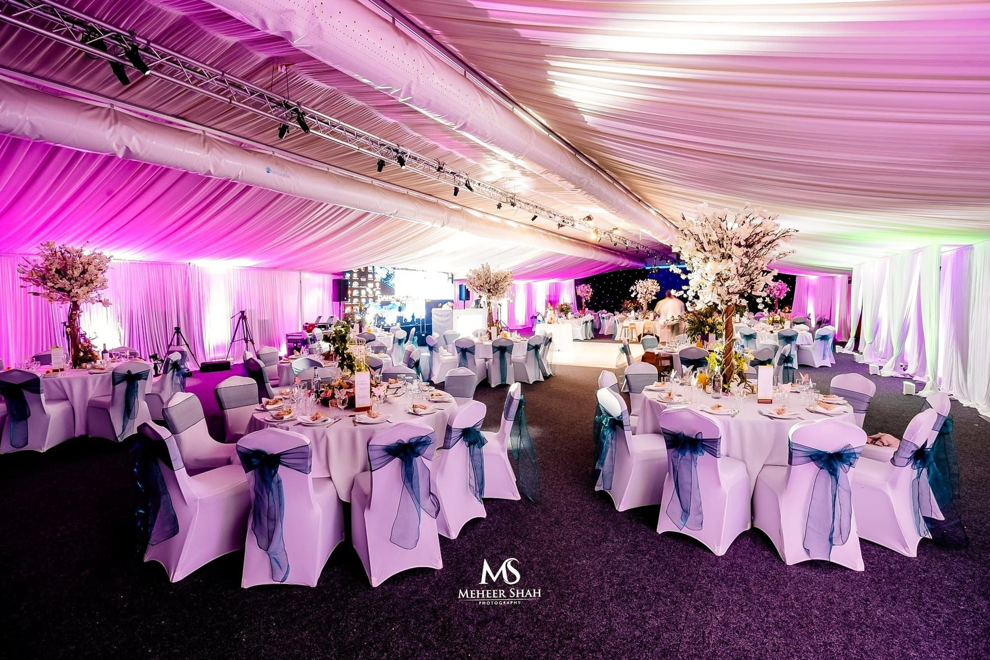 A marquee of round tables in white linen with organza chair sashes under pink uplighting