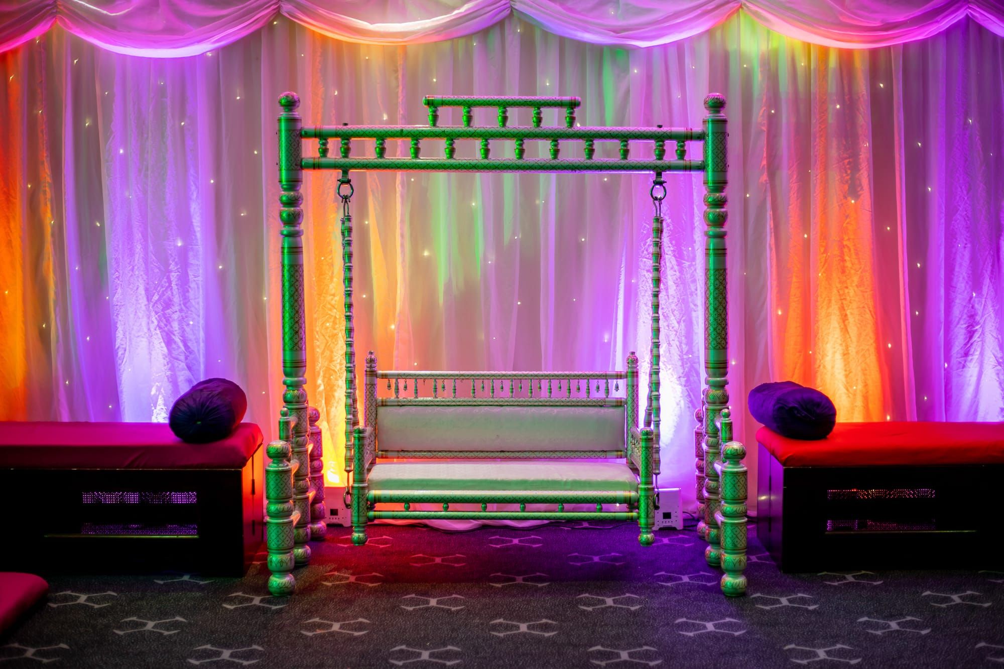 A green carved jhula swing set against drapes lit in orange, pink and purple