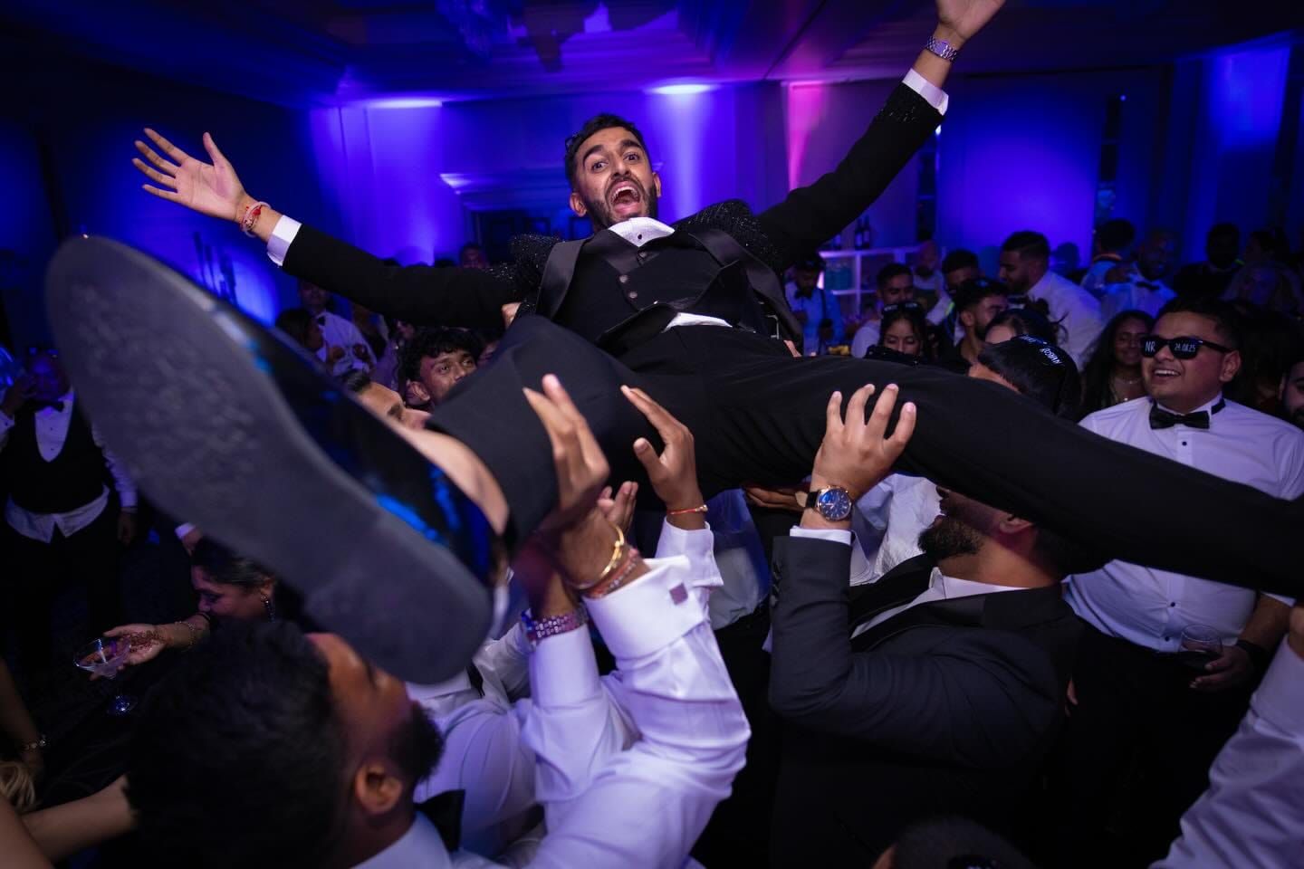 The groom lifted by guests on the dancefloor under blue and purple light