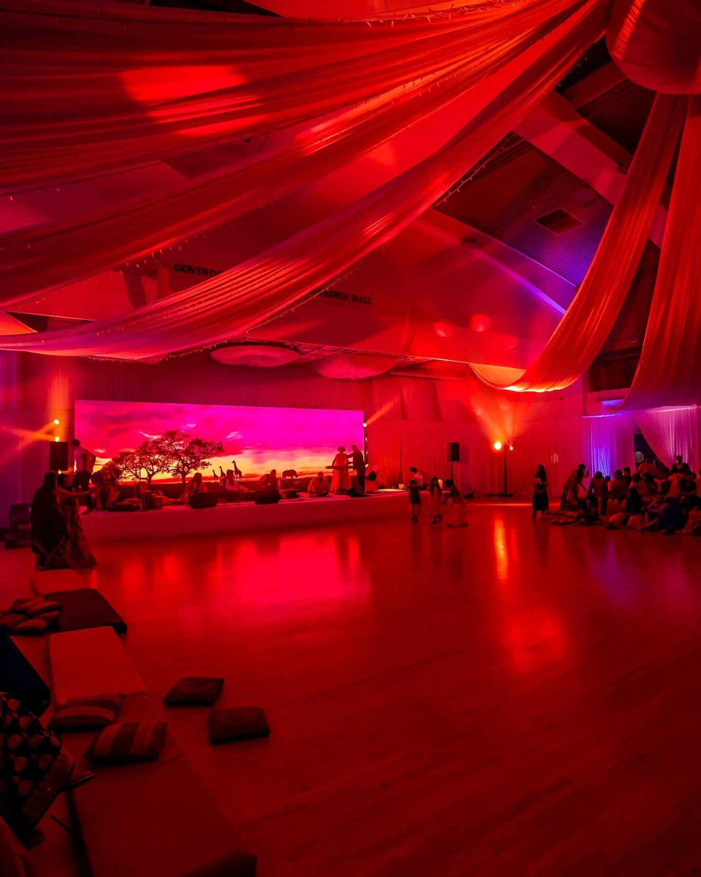 A hall draped and lit in deep red, guests seated along the far wall