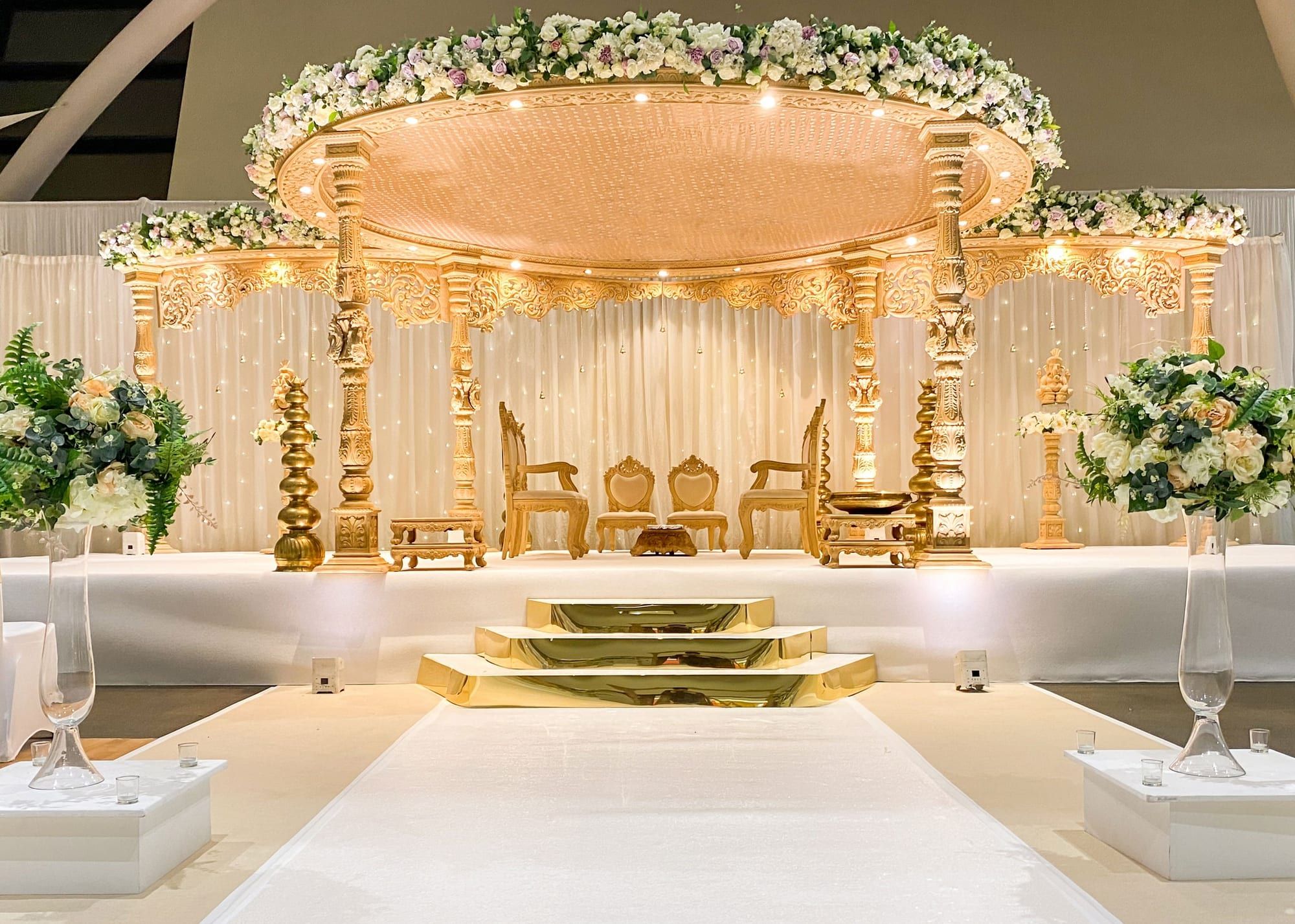A white and gold mandap dressed with florals, the aisle set for a ceremony