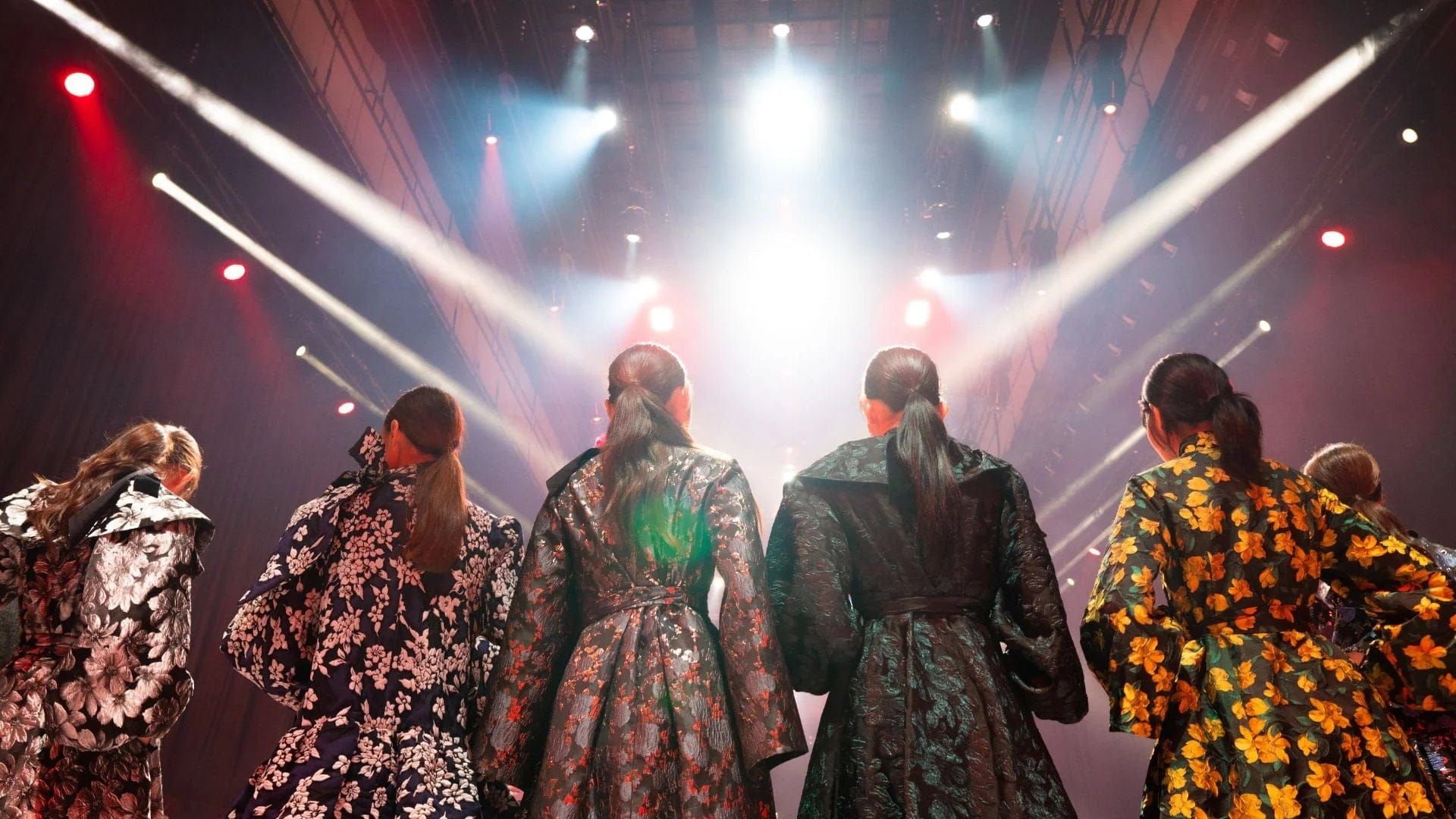 Models in printed gowns walking a runway under crossing spotlights