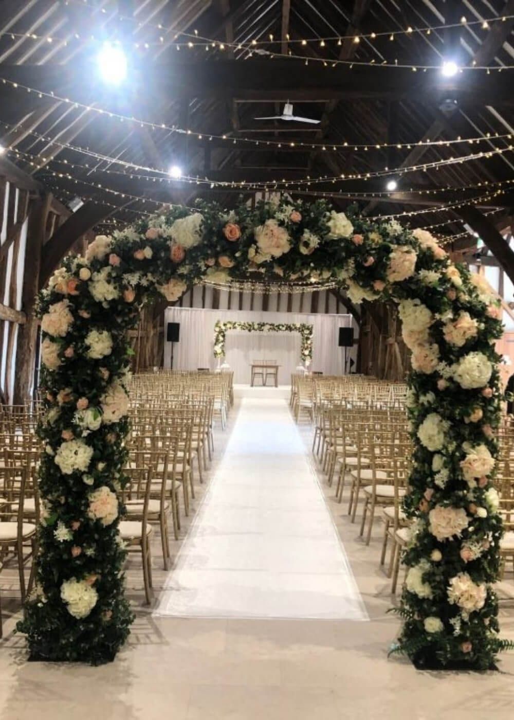 An arch of blush roses and foliage framing an aisle of chiavari chairs in a barn