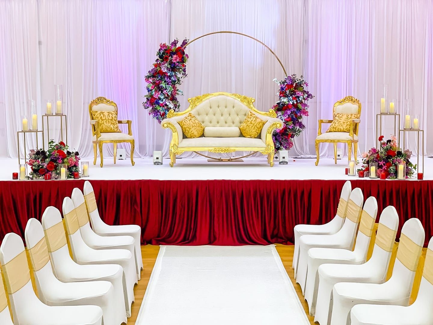 A gold sofa framed by a circular floral arch above a red-draped stage and white aisle