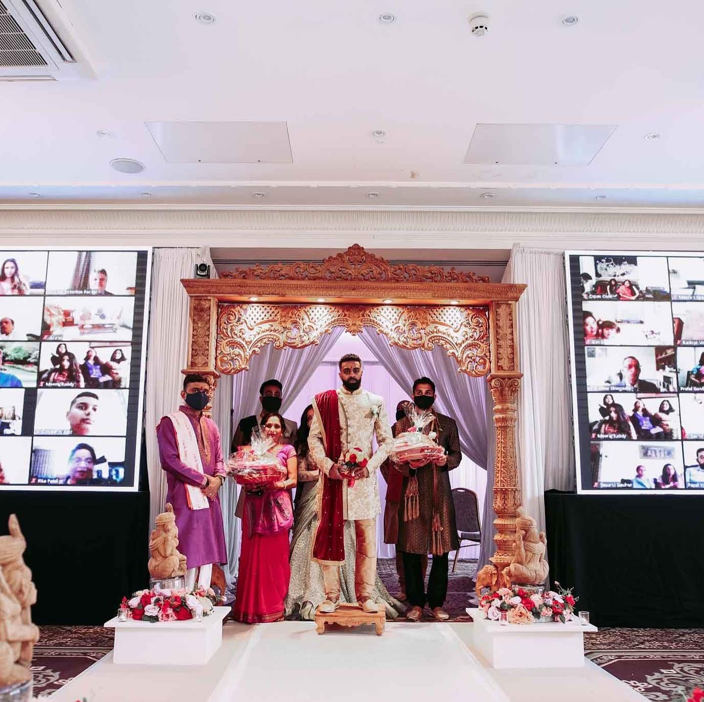 The couple at the mandap with two walls of video tiles showing family watching from home