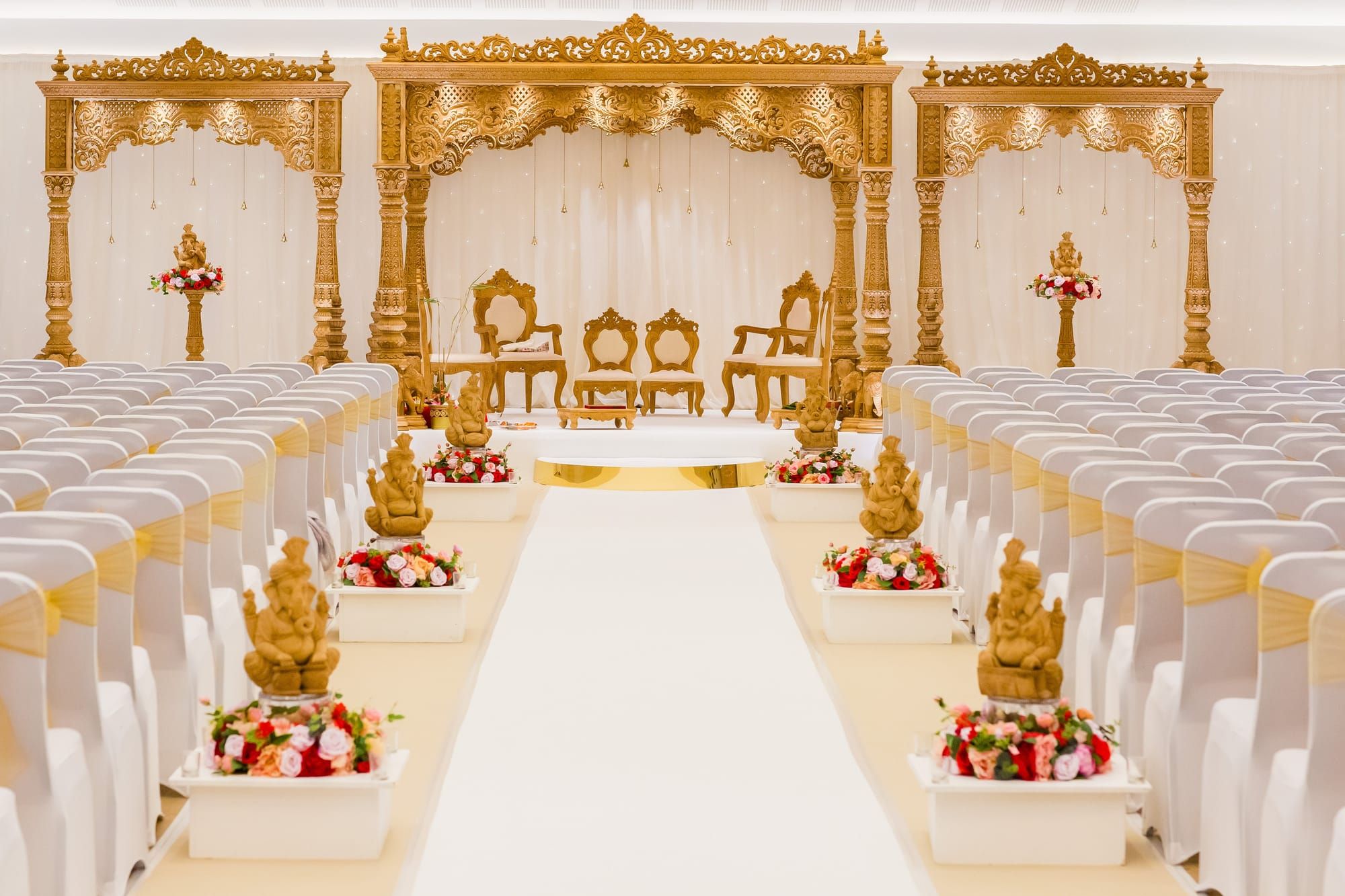 A hand-carved gold mandap with an aisle lined by Ganesh statues and floral trays