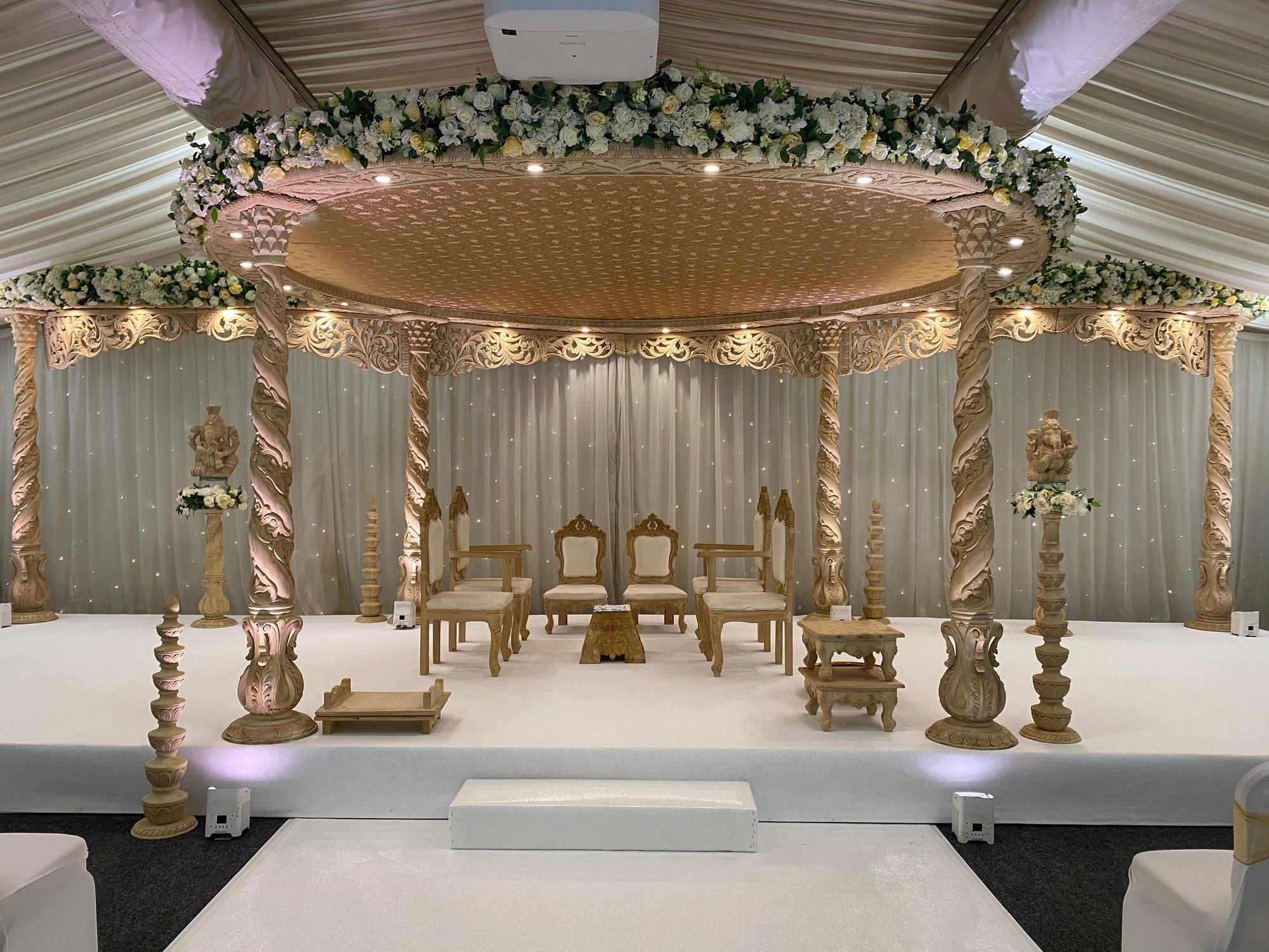 A carved mandap with a domed gold canopy on four pillars, set on a white stage before draped curtains