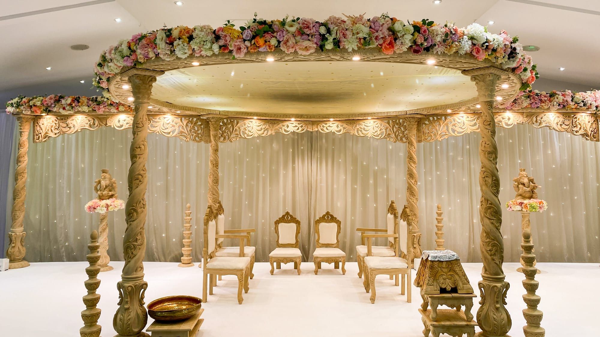 A gold mandap edged with pastel florals, its canopy lit from within by warm bulbs