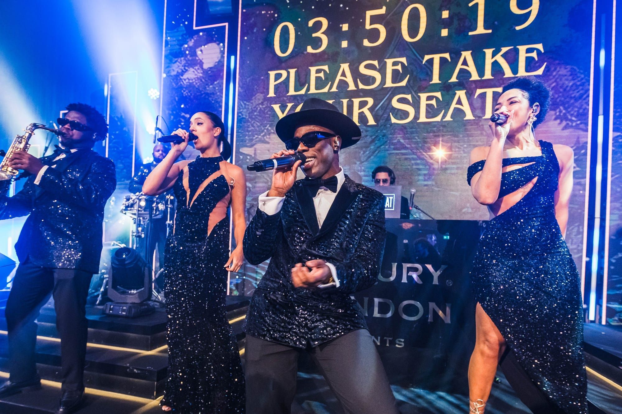 Three singers and a saxophonist performing together under stage lighting