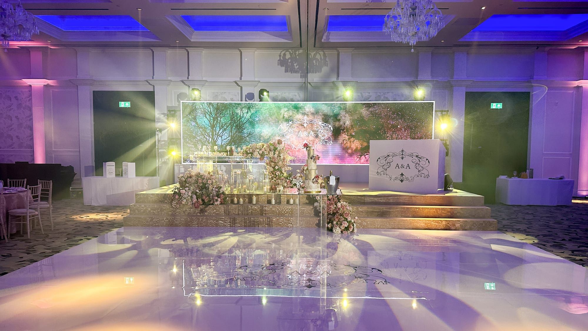 A ballroom stage framed by tall white floral arches above a mirrored dance floor under blue uplighting