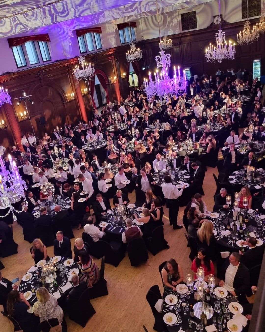 A black-tie dinner in full swing, hundreds of guests at round tables under chandeliers