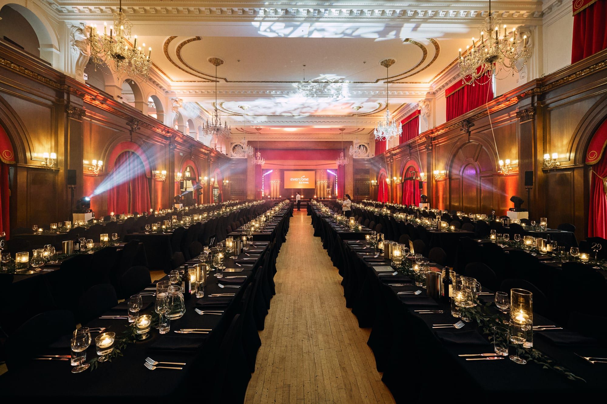 A ballroom set for a gala dinner, long candlelit banqueting tables running the length of the room under chandeliers and warm uplighting