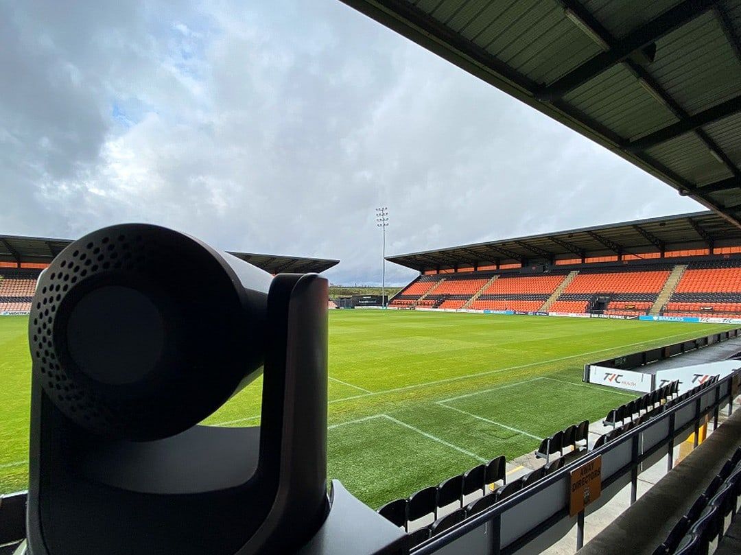 A broadcast camera on a gantry looking out over a stadium pitch and empty stand