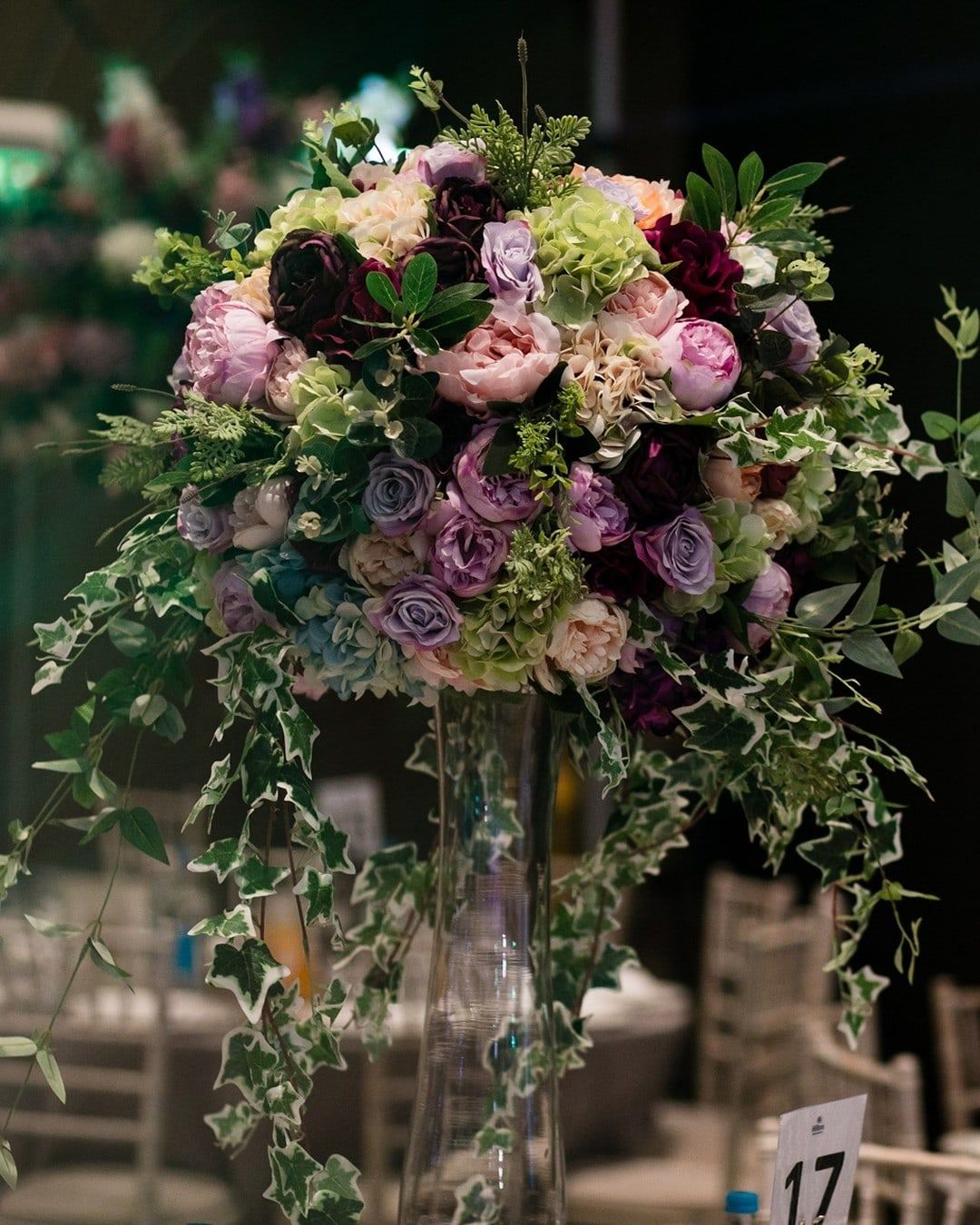 A tall arrangement of garden roses and trailing ivy on a glass stand above a laid table