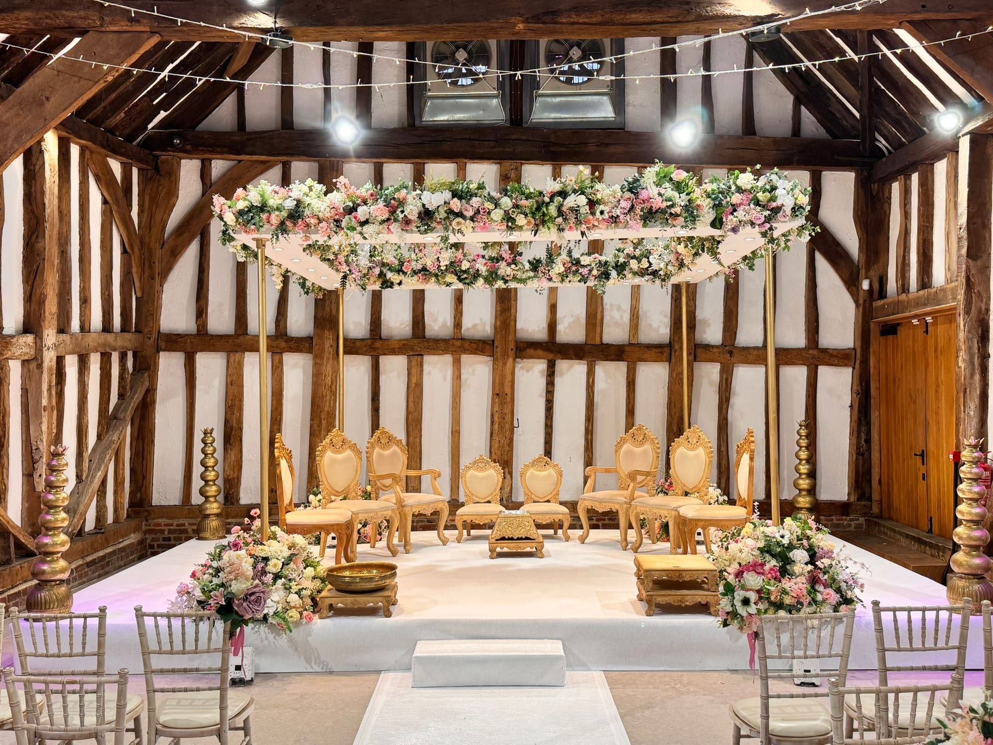 A mandap under the Great Barn's timber frame, a floral canopy strung between the beams