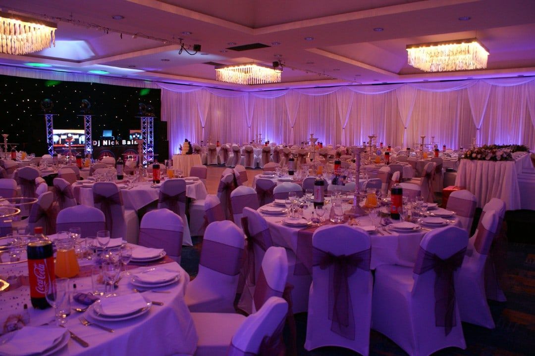 A banquet hall with draped walls washed in lilac uplighting, round tables laid beneath