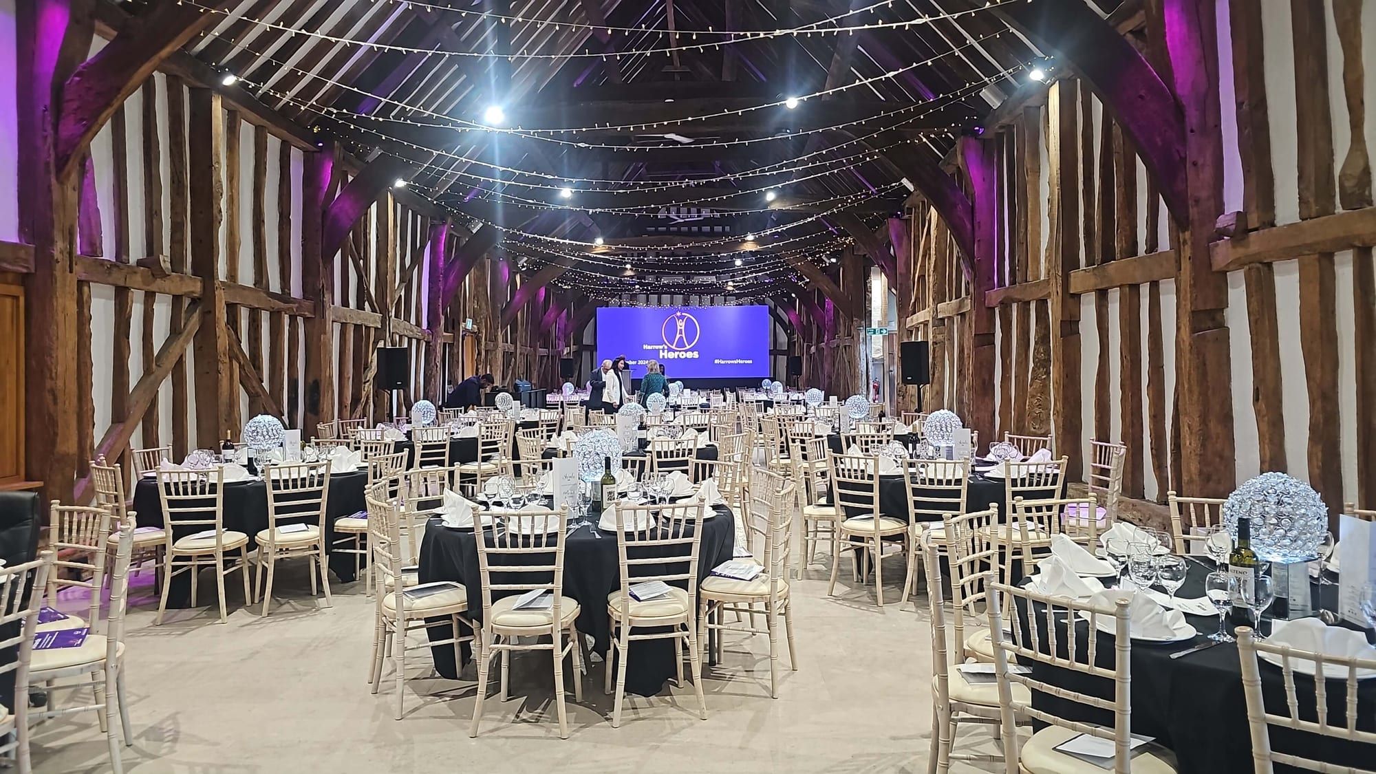 A timber-framed barn set for dinner, round tables under festoon lighting with a lit stage at the far end