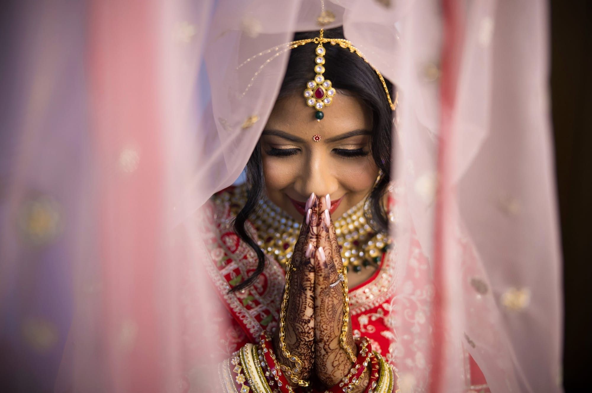 A bride beneath her veil with hands joined in greeting